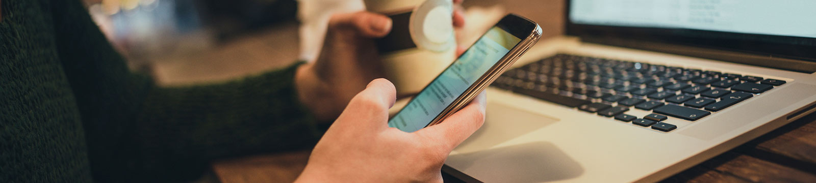 Hands holding phone and coffee with laptop on table.